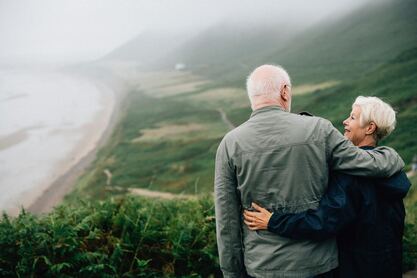 An elderly couple stands upon a cliff looking at each other. The scenery depicts a green overcast landscape with sand and ocean to the right. The woman's face is in view and she is smiling.
